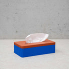 De Wavy Tissuebox Blauw/Terracotta van Wavy Design Studio, met een blauwe basis en terracotta deksel met een tissue gedeeltelijk uitgetrokken, rust op een licht oppervlak tegen een grijs gespikkelde muur.