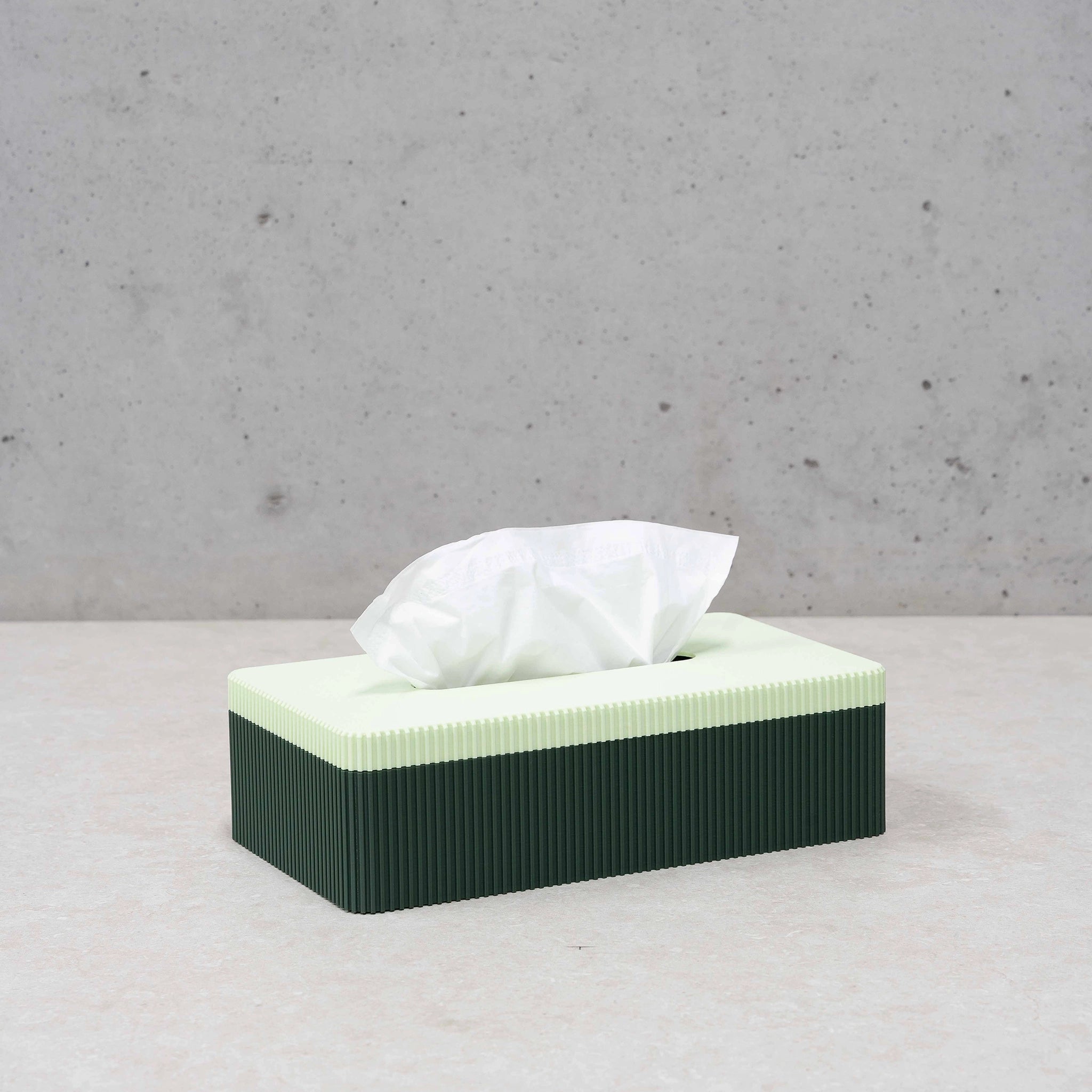 De Wavy Tissuebox Highland Groen/Munt van Wavy Design Studio staat op een lichte ondergrond met een wit tissue dat uit de lichtgroene deksel steekt. Op de achtergrond is een effen, lichtgrijze muur zichtbaar.