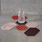 Een hoog glas bruisend water met ijs en fruit staat op een grijze tegel, omringd door zes hittebestendige Zone Denmark Triangles Trivet Kalamata siliconen onderzetters in rood-, roze- en bordeauxtinten.