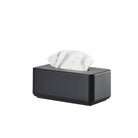 De Zone Denmark Ume Tissuebox Zwart, een strakke zwarte rechthoekige tissuebox van Zone Denmark met een wit tissue dat uit de bovenkant komt, benadrukt moderne elegantie tegen een effen witte achtergrond.