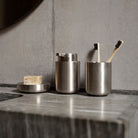 Drie roestvrijstalen badkameraccessoires van Zone Denmark, waaronder een zeepbakje, pompdispenser en de Ume Tandenborstelhouder met twee bamboetandenborstels, staan op een marmeren aanrecht tegen een betonnen muur.