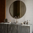 Een moderne badkamer met een ronde spiegel, grijze stenen wastafel, minimalistische kraan, zeep en de Zone Denmark Ume Tandenborstelhouder Staal op een betonnen aanrecht tegen een grijze muur met structuur.