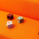 Drie kubusvormige Avolt USB-C stekkerdoos adapters, waaronder de Mulberry Rood editie geïnspireerd door Bauhaus Archiv, zitten op een fel oranje stoffen bank.
