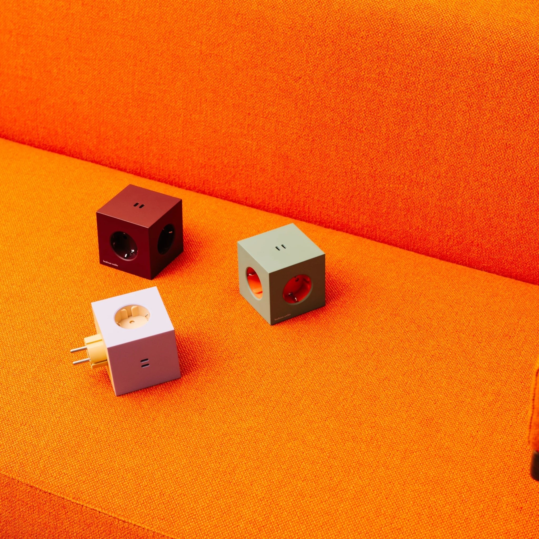 Drie kubusvormige Avolt USB-C stekkerdoos adapters, waaronder de Mulberry Rood editie geïnspireerd door Bauhaus Archiv, zitten op een fel oranje stoffen bank.