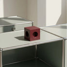 De Avolt x Bauhaus Archiv Square 2 USB-C Stekkerdoos Mulberry Rood van Avolt staat op een lichte metalen tafel met stalen buisranden, die een schaduw werpt in een heldere, minimalistische omgeving geïnspireerd op Bauhaus-design.