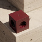 De Avolt x Bauhaus Archiv Square 2 USB-C Stekkerdoos in Mulberry Rood, een kubusvormige stroomadapter van Avolt, rust op beige stenen treden in het zonlicht.