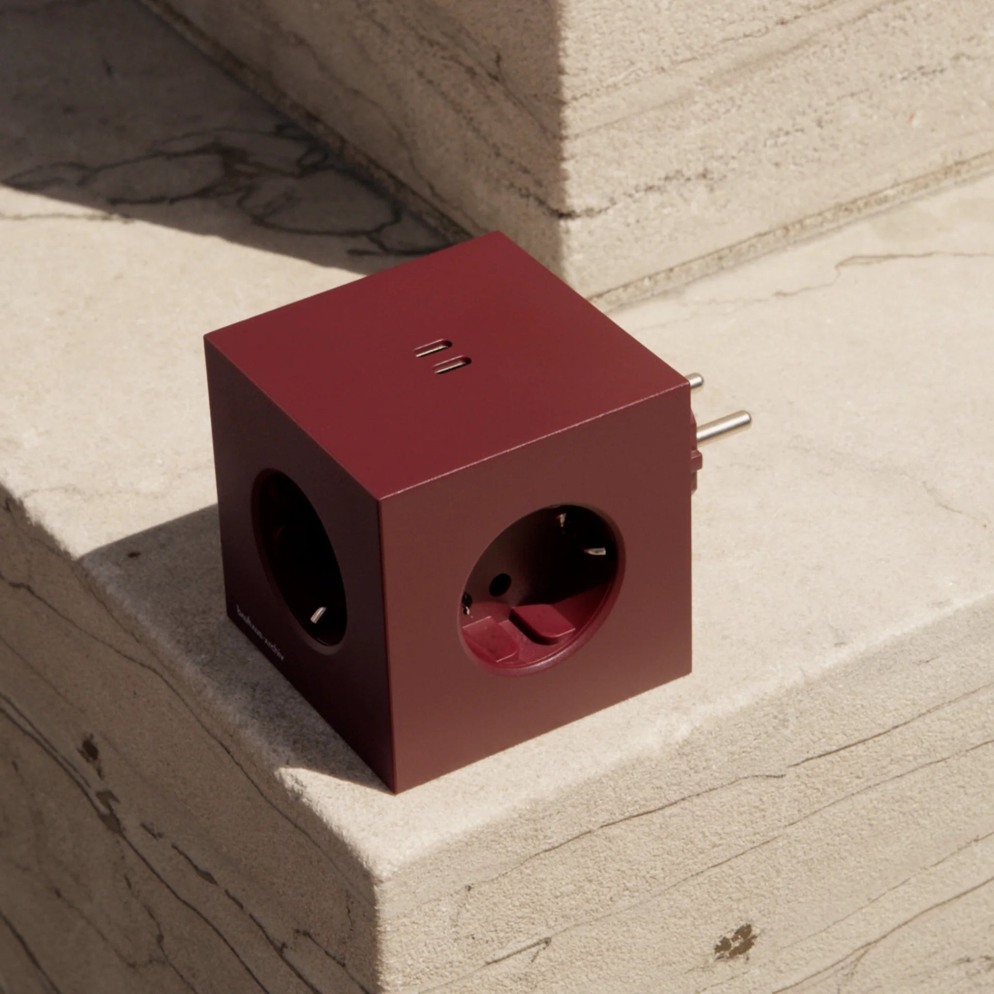 De Avolt x Bauhaus Archiv Square 2 USB-C Stekkerdoos in Mulberry Rood, een kubusvormige stroomadapter van Avolt, rust op beige stenen treden in het zonlicht.