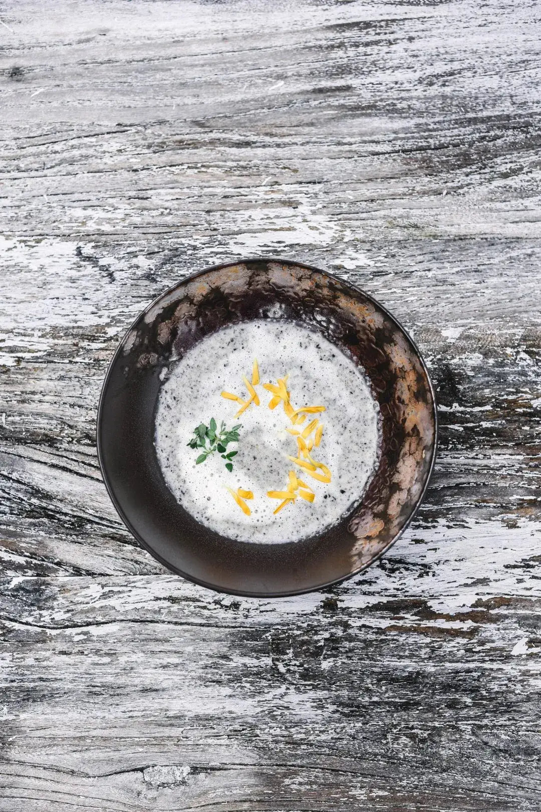 Een donkere kom romige soep geïnspireerd door Cozy Publishing Eat Finland Kookboek staat op een rustiek houten oppervlak, gegarneerd met geraspte kaas en een klein takje kruiden.