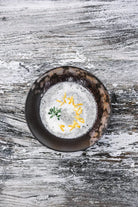 Een donkere kom romige soep geïnspireerd door Cozy Publishing Eat Finland Kookboek staat op een rustiek houten oppervlak, gegarneerd met geraspte kaas en een klein takje kruiden.