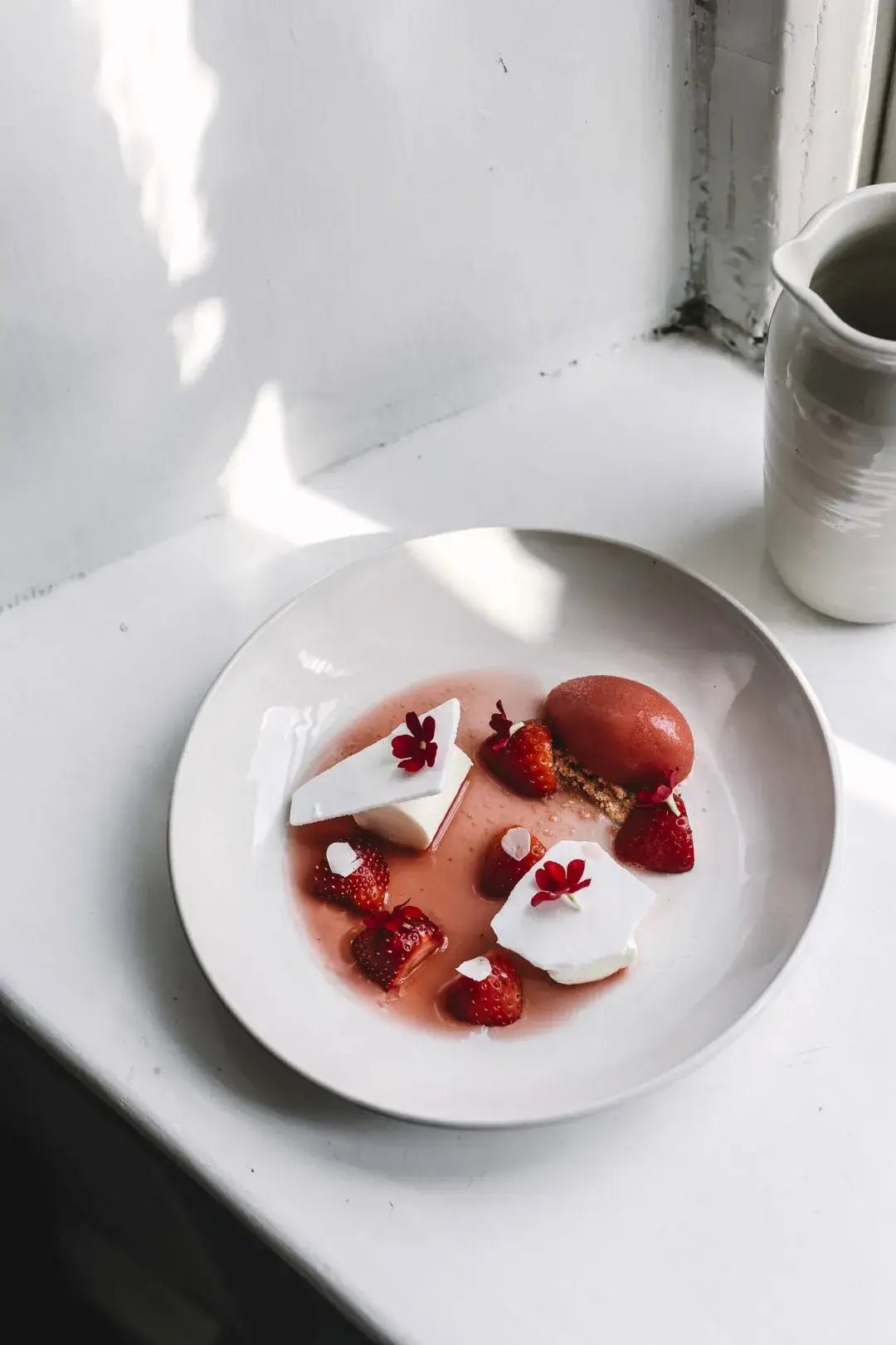 Een wit bord met witte meringue, verse aardbeien, rode saus, roze sorbet en eetbare rode bloemen staat op de vensterbank - een verleidelijke traktatie geïnspireerd op Finse recepten uit het Cozy Publishing Eet Finland Kookboek.