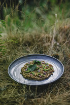 Een keramisch bord risotto gegarneerd met groen en paarse bloemen rust op droog gras, wat de Scandinavische sfeer oproept die te vinden is in het Eet Finland Kookboek van Cozy Publishing.