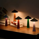 Vier compacte, kegelvormige Fine Lumens Tipsy Lamp LED Oplaadbare tafellampen in Poederroze en Lichtpaars verlichten zacht een houten tafel met boeken. Warm licht gloeit terwijl een potplant op de achtergrond verschijnt.