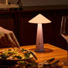 De Fine Lumens Tipsy Lamp LED Oplaadbaar Poederroze Lichtpaars brandt warm op een houten eettafel naast iemand die salade eet. Een wijnglas en wat eten maken deze zacht verlichte scène van Fine Lumens extra gezellig.