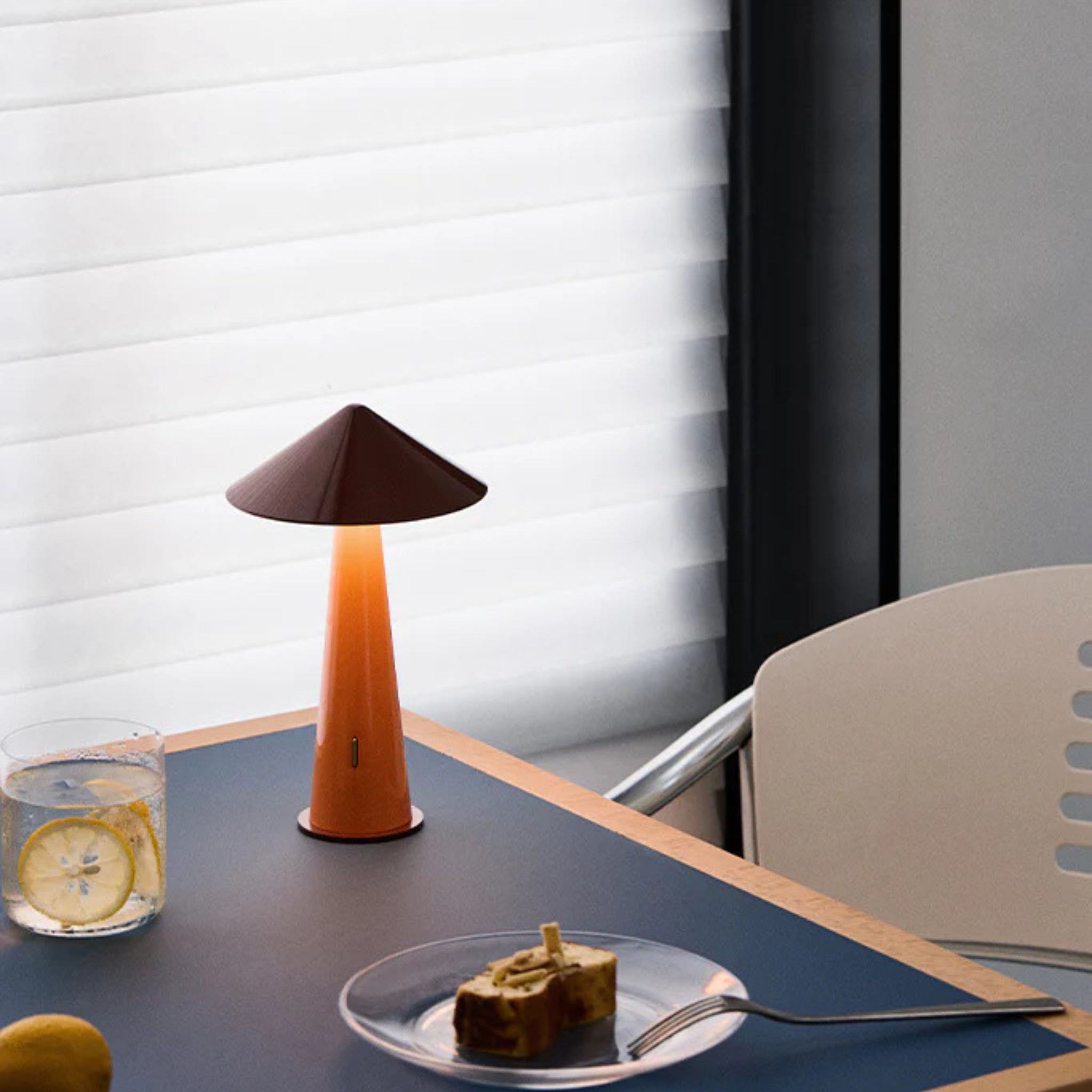 Op een moderne tafel met een blauw oppervlak liggen een Fine Lumens Tipsy Lamp LED Oplaadbaar Rustiek Rood Oranje, een sinaasappel, een glas water met citroen, een vork en een bord met gebak. Achter de tafel staat een witte stoel en een raam met witte jaloezieën.