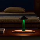 De Fine Lumens Tipsy Lamp LED Oplaadbaar Zwart Emerald Groen gloeit zacht op een houten nachtkastje voor een keurig opgemaakt bed met bruin en grijs beddengoed in een schemerige slaapkamer.