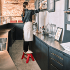 Een vrouw in een schort staat op een Hasegawa Lucano Ladder 1 Step Rood, reikend naar een plank in een moderne keuken met donkere kastjes, flessen, potten en een ingelijste menukaart op het aanrecht.