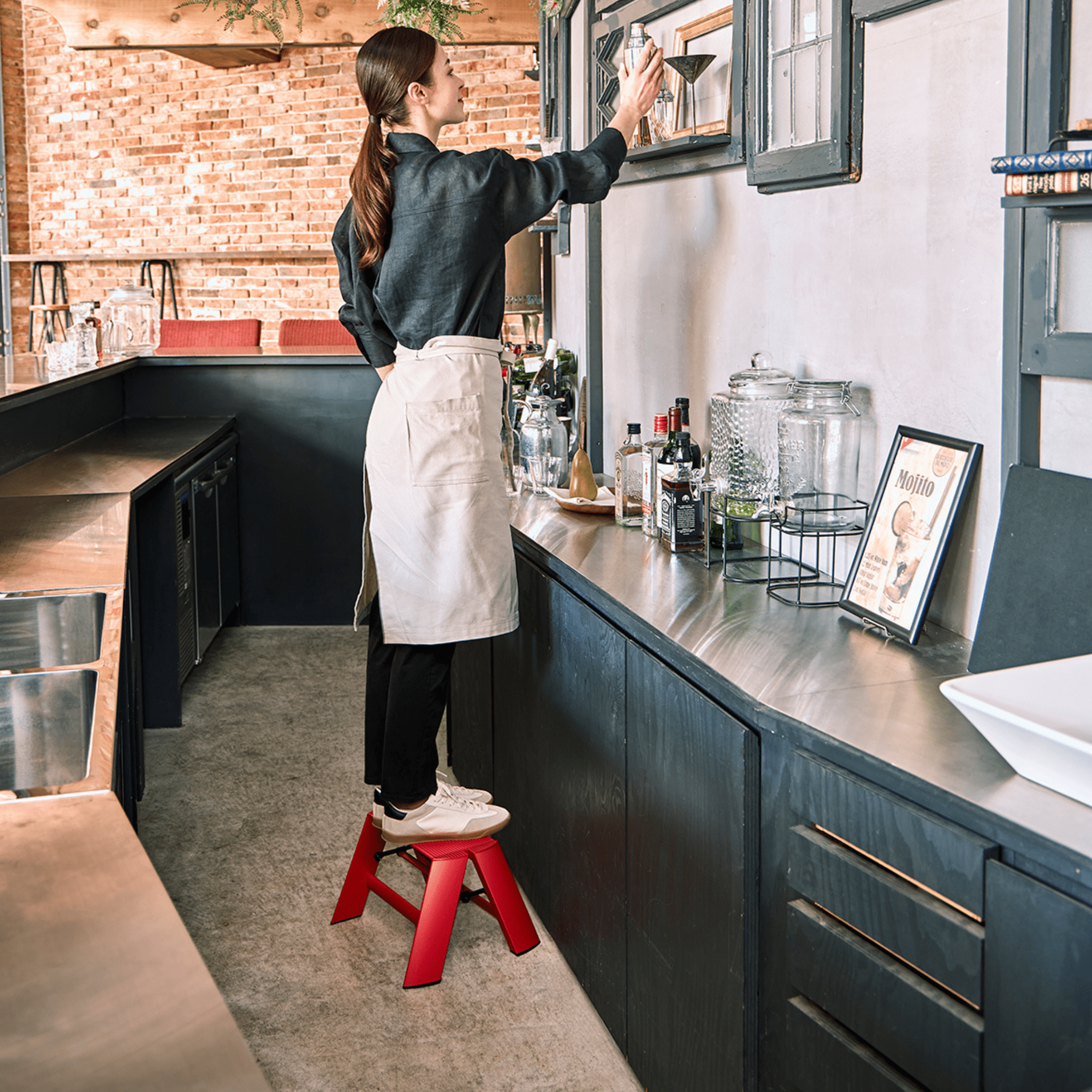Een vrouw in een schort staat op een Hasegawa Lucano Ladder 1 Step Rood, reikend naar een plank in een moderne keuken met donkere kastjes, flessen, potten en een ingelijste menukaart op het aanrecht.