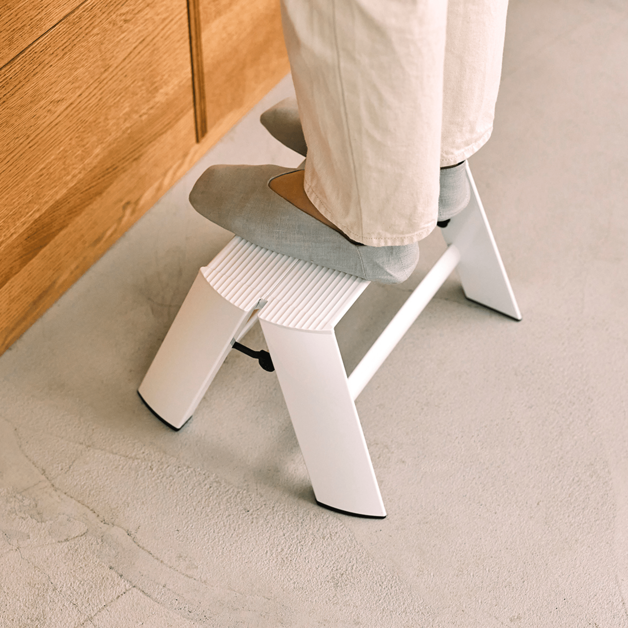 Een persoon in een lichtgekleurde broek en grijze schoenen staat op een Hasegawa Lucano Ladder 1 Step Wit, een compact wit opstapkrukje van Hasegawa met Japans design, geplaatst op een lichtgrijze vloer naast een houten kast.