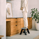 Een persoon in lichte kleding gebruikt de Hasegawa Lucano Ladder 1 Trede Zwart van Hasegawa om bij een houten kast te komen in een moderne, Japans geïnspireerde woning. Een potplant en ingelijste abstracte kunst versieren de ruimte.