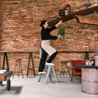 Staande op een Hasegawa Lucano Ladder 2 Stap Dusty Mint, schikt een persoon een hangende plant op een plank in een kamer met zichtbare bakstenen, barkrukken, een rode stoel en rustgevende dusty mint decoraccenten.