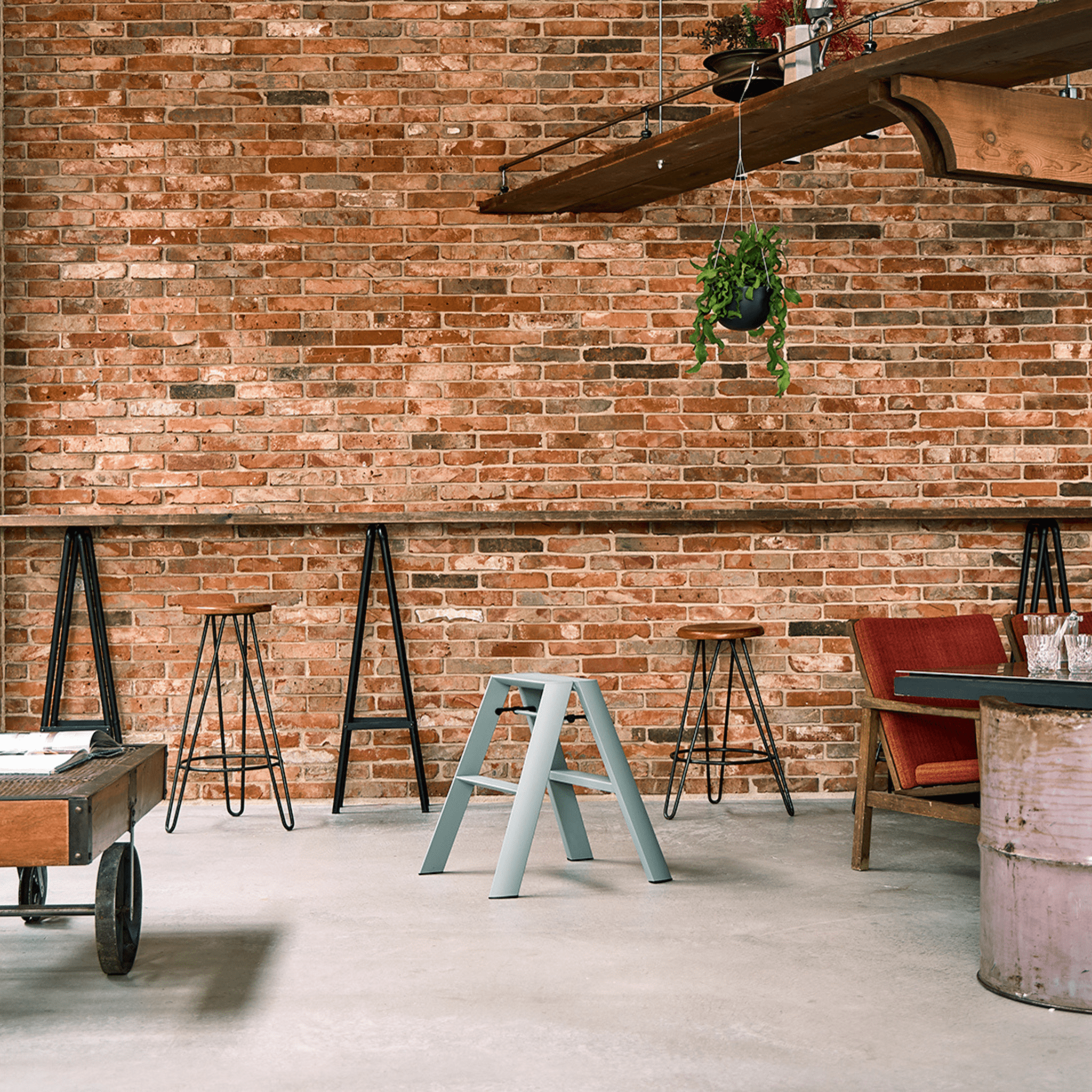 Een kamer in loftstijl met een bakstenen muur, verschillende krukjes, een Hasegawa Lucano Ladder 2 Step Dusty Mint, een rode stoel met kussens, een houten tafel en een hangende plant - allemaal op een betonnen vloer en geaccentueerd met industriële decoraties.