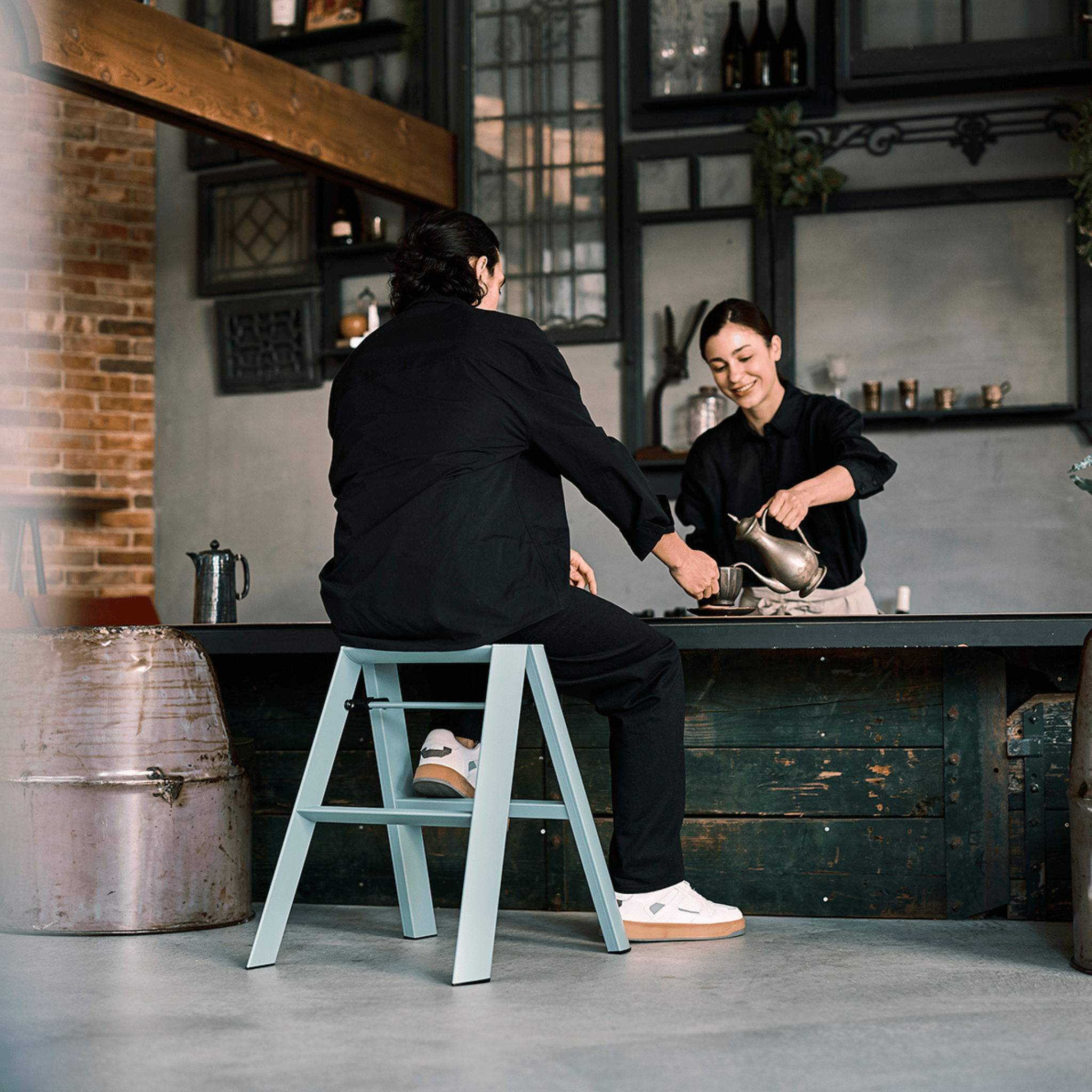 Een persoon zit op een Hasegawa Lucano Ladder 2 Step Dusty Mint aan een toonbank terwijl een ander een drankje inschenkt. Beiden glimlachen in een gezellig café met zichtbare bakstenen en donkerhouten accenten.