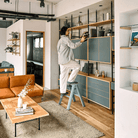 Staande op een Hasegawa Lucano Ladder 2 Step Dusty Mint, plaatst een persoon decor op een hoge plank in een moderne woonkamer met Japans design, blauwe kasten en houten vloeren.