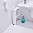 De Hasegawa Lucano Ladder 2 Step Wit van Hasegawa, een compacte witte vouwladder geïnspireerd op Japan, staat tegen een betegelde badkamermuur bij een wastafel; een groene glazen fles staat op de vloer en een handdoek hangt erboven.