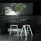 Een minimalistische kamer heeft een donkere rechthoekige tafel met een hoge glazen vaas met groene takken, glaswerk en een karaf. Op de schemerige achtergrond roepen twee Hasegawa Lucano Ladder 2 Step Wit krukken een modern Japans geïnspireerd ontwerp op.