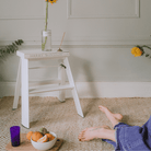 Een gezellige scène met de Hasegawa Lucano Ladder 2 Step Wit van Hasegawa met een vaas met een gele bloem en boeken, met fruit op een dienblad en een paars glas in de buurt, terwijl iemand op blote voeten in een blauwe spijkerbroek zit.