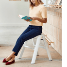 Een vrouw in een lichtgele top en blauwe jeans zit op een Hasegawa Lucano Ladder 2 Step Wit van Hasegawa en leest een turquoise boek. Ze draagt rode platte schoenen binnenshuis bij een toonbank met boeken en decor, die subtiele Japanse designelementen weerspiegelen.