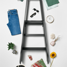 De Hasegawa Lucano Ladder 3 Step Zwart van Hasegawa staat rechtop op een witte achtergrond, met voorwerpen zoals blauwe jeans, een groene handdoek, zonnebrillen, een klok, een plant, een zonnebloem, mokken, schelpen en een gestreepte mat eromheen verspreid.