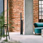 Een minimalistische leefruimte met zichtbare bakstenen, een groenblauwe fauteuil en een vleugje Japans design. Een moderne flatscreentelevisie staat bij de betonnen pilaar, terwijl een Hasegawa Lucano Ladder 3 Step Zwart van Hasegawa gedeeltelijk zichtbaar is aan de linkerkant. Zonlicht valt naar binnen door de grote ramen.