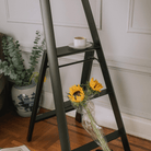 Een boeket zonnebloemen ligt bovenop de Hasegawa Lucano Ladder 3 Step Zwart van Hasegawa, die ook een kop en schotel bevat. Een potplant staat vlakbij op een houten vloer, tegen een Japandi-stijl muur met witte panelen.