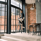 Een persoon staat op een Hasegawa Lucano Ladder 4 Stap Zwart van Hasegawa in een moderne industriële kamer met grote ramen, bakstenen muren en zwart meubilair, papieren vasthoudend en naar beneden kijkend terwijl hij schoonmaakt.