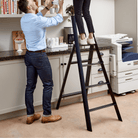 Een man staat naast een vrouw die de Hasegawa Lucano Ladder 4 Step Zwart van Hasegawa gebruikt om een hoge keukenplank te bereiken in een kantoorruimte met kasten, aanrecht en een printer op de achtergrond.