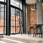 Een moderne ruimte in industriële stijl met grote glazen ramen, zichtbare bakstenen muren, de Hasegawa Lucano Ladder 4 Step Zwart, zwarte krukjes, een ronde tafel en een hangende plafondlamp badend in natuurlijk licht.