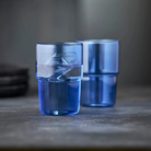 Twee Lyngby Glas Torino glazen blauw 40 cl zijn te zien: één op de voorgrond gevuld met ijs en water, de ander leeg en vervaagd op de achtergrond. Beide van stevig borosilicaatglas, tegen een donker oppervlak met grijze achtergrond.