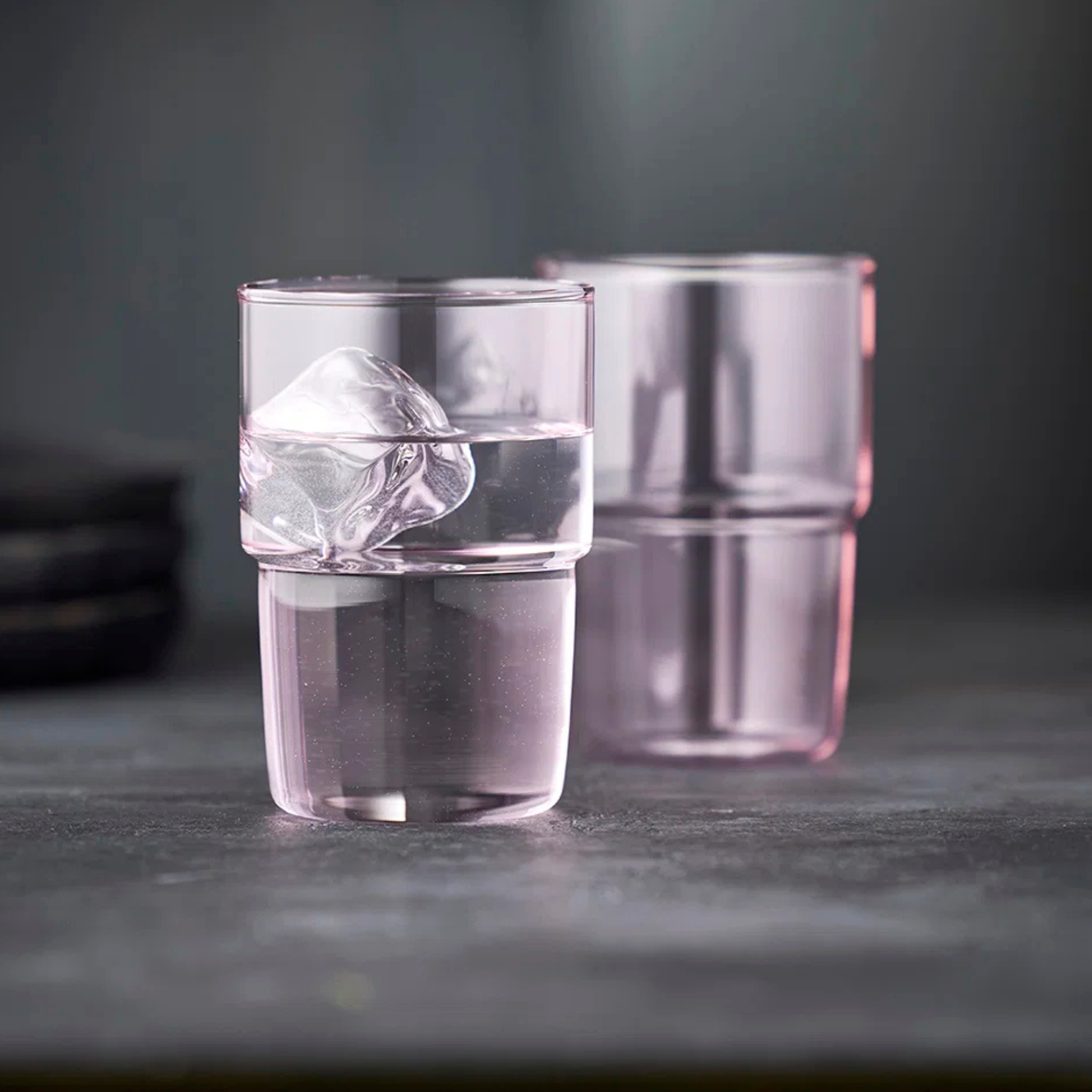 Twee Lyngby Glas Torino Roze 40 cl glazen zijn gestapeld, de bovenkant gevuld met water en ijs. De vage grijze achtergrond benadrukt de minimalistische, moderne stijl van deze stijlvolle Lyngby Glas drinkglazen.