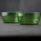 Twee kleine, ronde Lyngby Glas Torino Kom Groen D12 kommen (set van 2) van Lyngby Glas rusten op een grijs oppervlak met een effen donkergrijze achtergrond. De voorste kom is scherp gefocusseerd; de achterste kom is een beetje wazig.