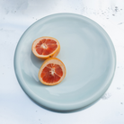 Twee helften van een grapefruit liggen met de snijkant naar boven op een Matias Moellenbach Big O Serveerschaal in de delicate kleur Blue Haze, die op een wit oppervlak met subtiele sporen en vlekjes staat.
