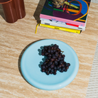 Op de Matias Moellenbach Big O-serveerschaal in Blue Haze, die op een beige stenen tafel staat, ligt een tros donkere druiven, met daarnaast een stapel kleurrijke tijdschriften en een bruine vaas.