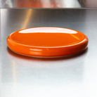 De Matias Moellenbach Big O Serveerschaal Orange, een glanzende, platte porseleinen serveerschaal van Matias Moellenbach, staat op een spiegelend metalen oppervlak met een metalen wand op de achtergrond.