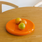 Drie sinaasappels liggen op een „Big O Serveerschaal Orange“ van Matias Moellenbach, die op een lichte houten tafel staat, met op de achtergrond een houten stoel met een gebogen rugleuning.