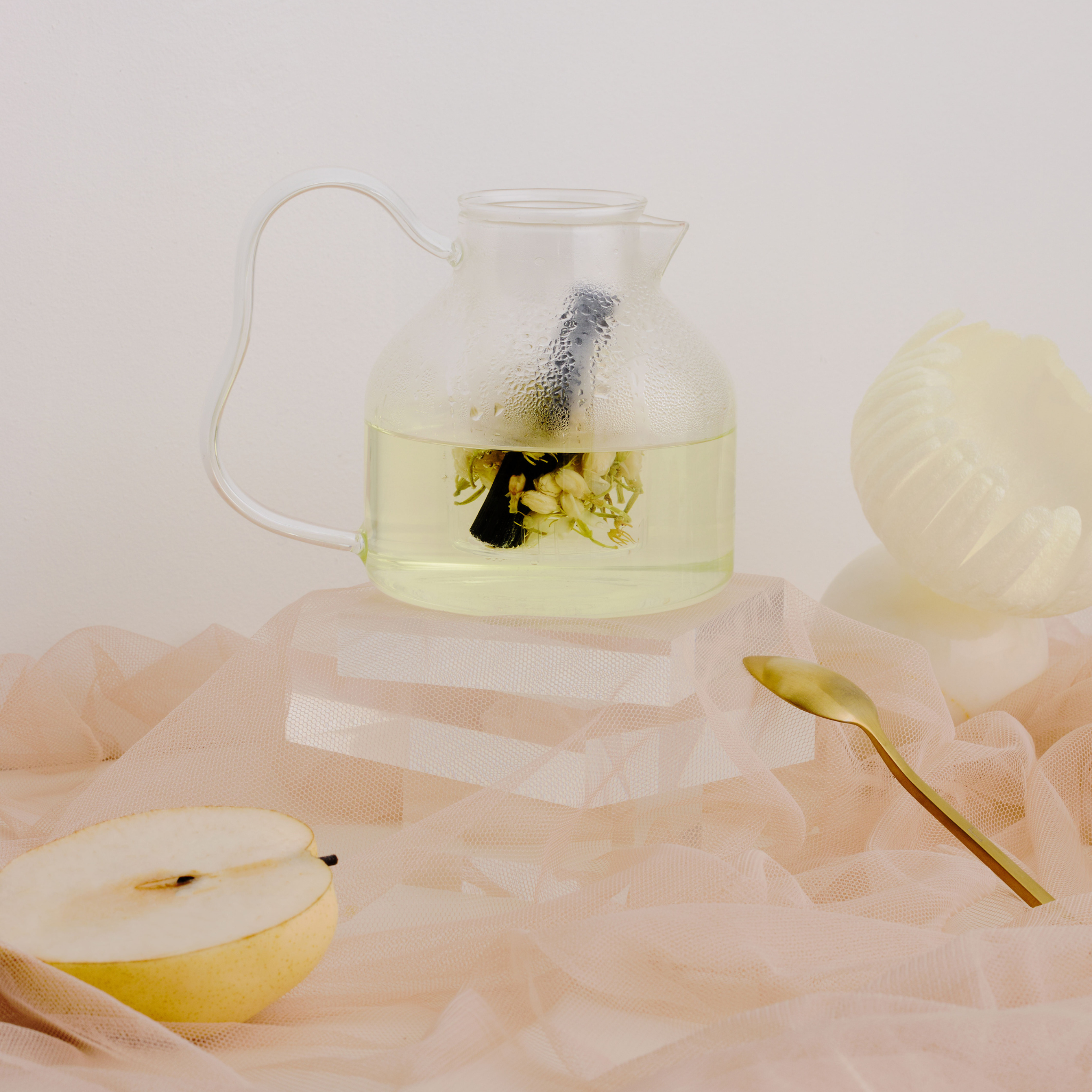 Een Matias Moellenbach Bincho-theepot van Matias Moellenbach, vervaardigd uit hittebestendig glas, is gevuld met bloementhee en wordt vergezeld door een gouden lepel, een geschilde peer en een halve peer – allemaal kunstzinnig gepresenteerd op zachte beige stof tegen een minimalistische, elegante achtergrond.