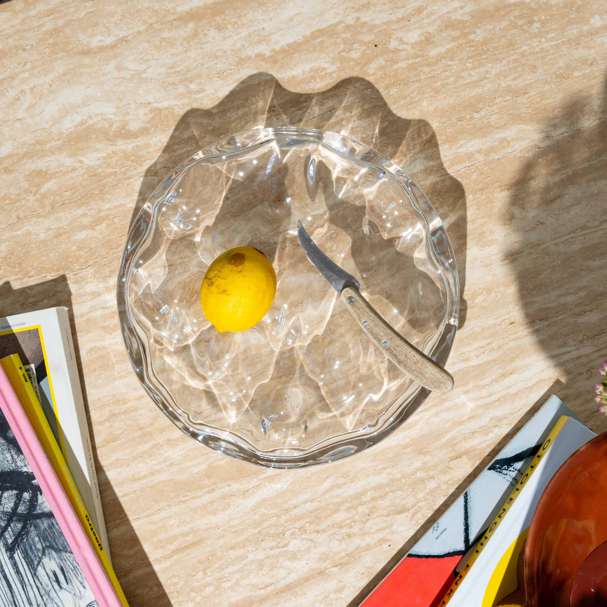 De Matias Moellenbach Blister Bowl, vervaardigd uit helder glas, bevat één citroen en een klein mesje en staat op een beige marmeren oppervlak naast een aantal kleurrijke tijdschriften, waardoor dit alledaagse tafereel wordt omgetoverd tot een opvallend designobject van Matias Moellenbach.