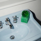 Een mintgroene Matias Moellenbach OBA Zeep + Houder Aloe Vera-zeep, met daarbovenop een ronde groene bol, ligt op een witte wastafel naast zilveren kraanhendels, met hier en daar wat waterdruppels – een toonbeeld van de subtiele Scandinavische designelegantie van Matias Moellenbach.