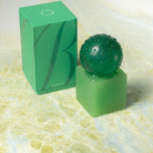 Een groene doos, versierd met een gestileerde B, staat naast een bijpassende groene kubus met daarop een groene bol met structuur; beide staan op een licht gemarmerd oppervlak. Het tafereel belichaamt de strakke esthetiek van Scandinavisch design, waardoor het een ideale presentatie vormt voor de Matias Moellenbach OBA Zeep + Houder Aloe Vera van Matias Moellenbach.