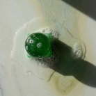 Een groen ijsblokje met belletjes op het oppervlak smelt op een lichtgekleurde ondergrond, wat de frisse, levendige uitstraling van de Matias Moellenbach OBA Zeep + Houder Aloe Vera van Matias Moellenbach weerspiegelt. Terwijl zich eronder een klein plasje vormt en een diagonale schaduw een deel van het tafereel siert, roept de compositie door haar eenvoud en rustige esthetiek elementen van Scandinavisch design op.