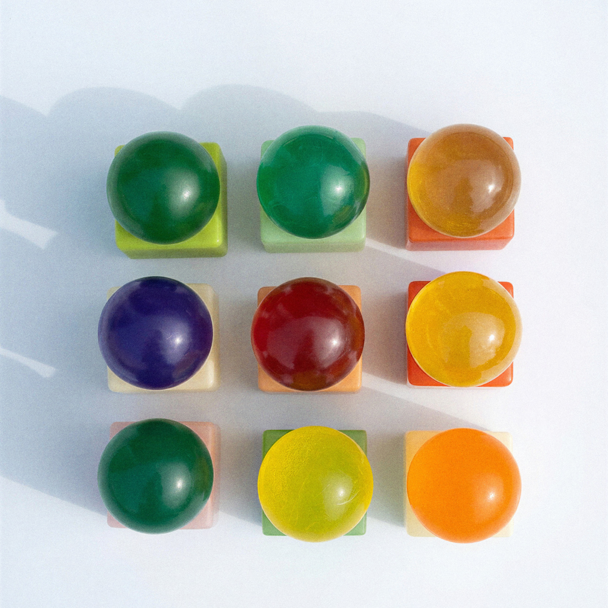Negen levendige, doorschijnende bollen, elk geplaatst op een vierkante pastelkleurige voet en gerangschikt in een raster van 3 bij 3, weerspiegelen de speelse esthetiek van de Matias Moellenbach OBA Zeep Bar Aloe Vera. Elke bol heeft een unieke tint – waaronder groen, geel, oranje, rood en paars – die aansluit bij de levendige kleuren van de vaste zeep van Matias Moellenbach en zachte schaduwen werpt op een witte achtergrond.